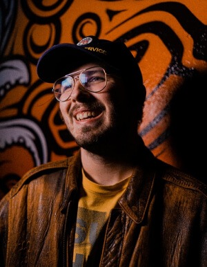 A smiling person with glasses wearing a baseball cap and leather jacket standing in front of an orange graffiti wall