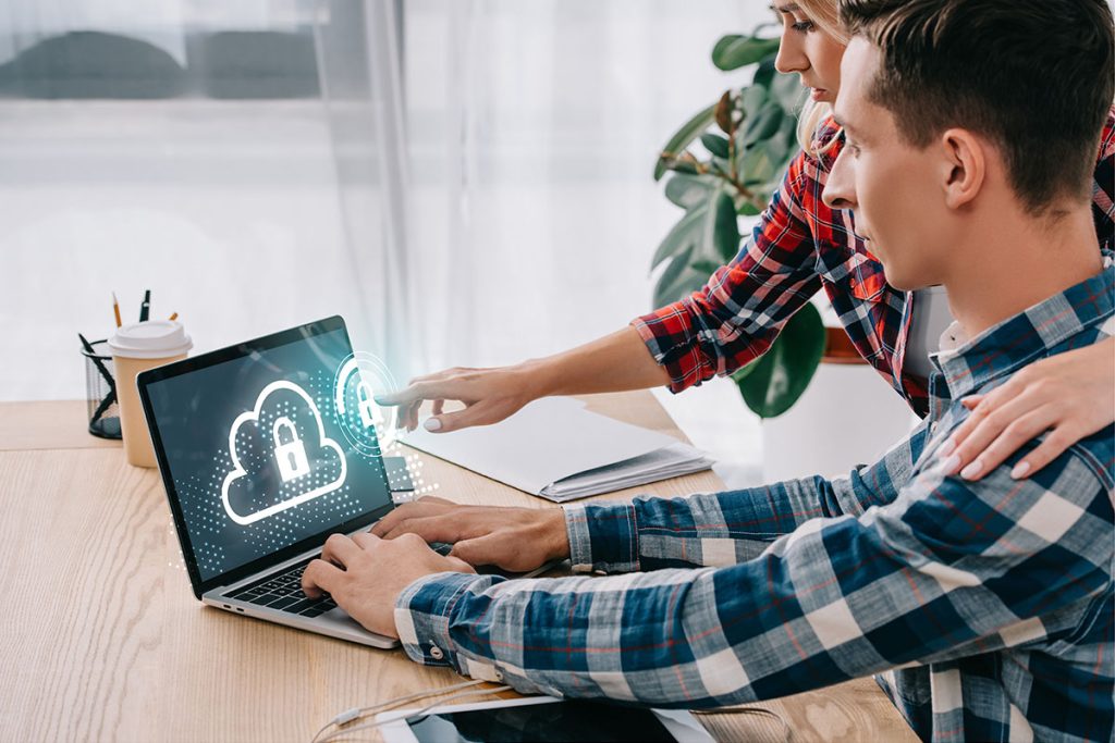 A man and a woman are interacting with a laptop on a wooden table, with digital cloud and lock icons displayed on the screen, symbolizing cybersecurity. The woman is pointing at the screen while placing her hand on the man's shoulder. A coffee cup and stationery are visible in the background.