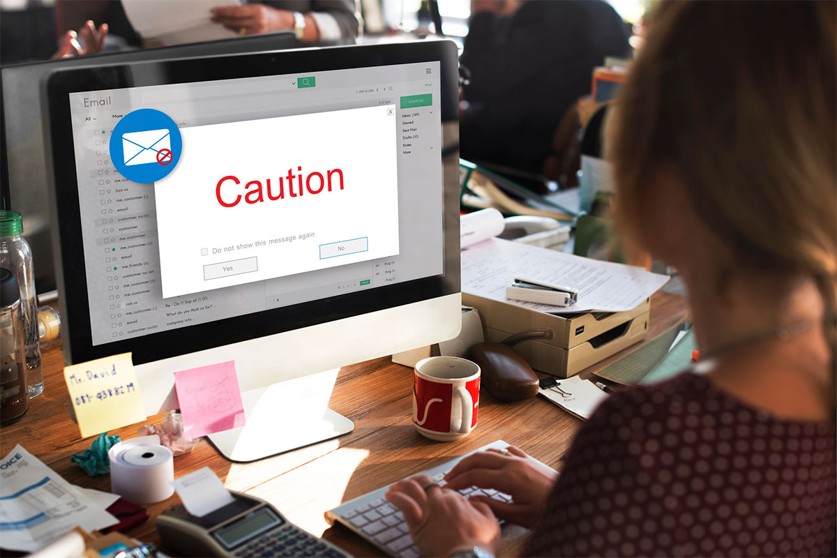 Email Caution A person is typing on a keyboard in front of a computer displaying an email warning message that says "Caution" in red. The desk is cluttered with paperwork, a red mug, and various office supplies.
