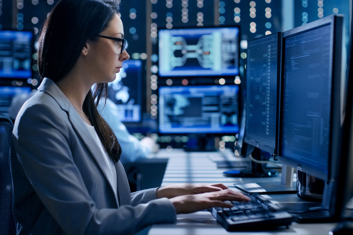 Managed IT Concept A woman in a gray blazer and glasses types on a keyboard in a dimly lit room filled with multiple computer monitors displaying data and graphs.