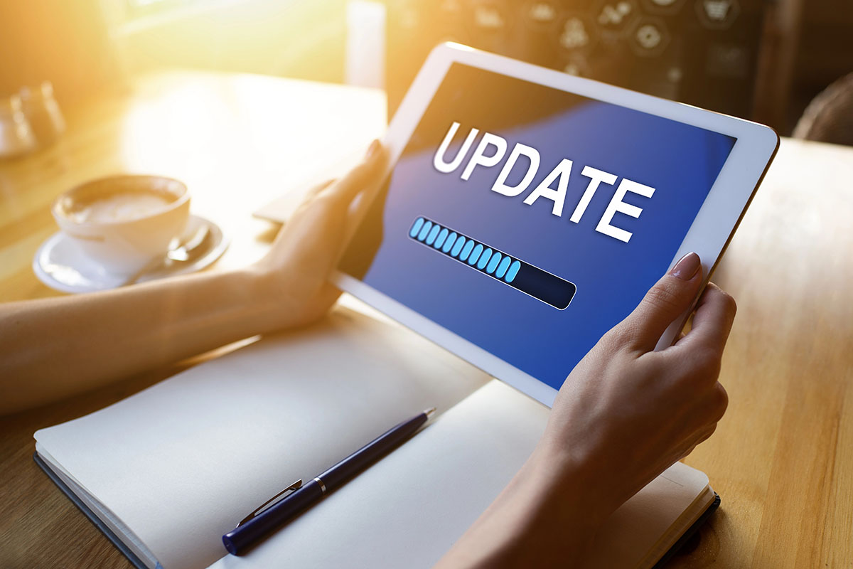Security Update Concept A person holding a tablet displaying an "UPDATE" screen with a progress bar, next to a notebook and a cup of coffee on a wooden table, illuminated by sunlight.