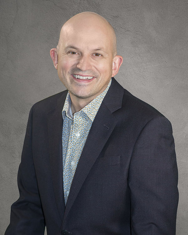 A smiling bald man wearing a dark blazer over a patterned shirt, posed against a textured gray background.