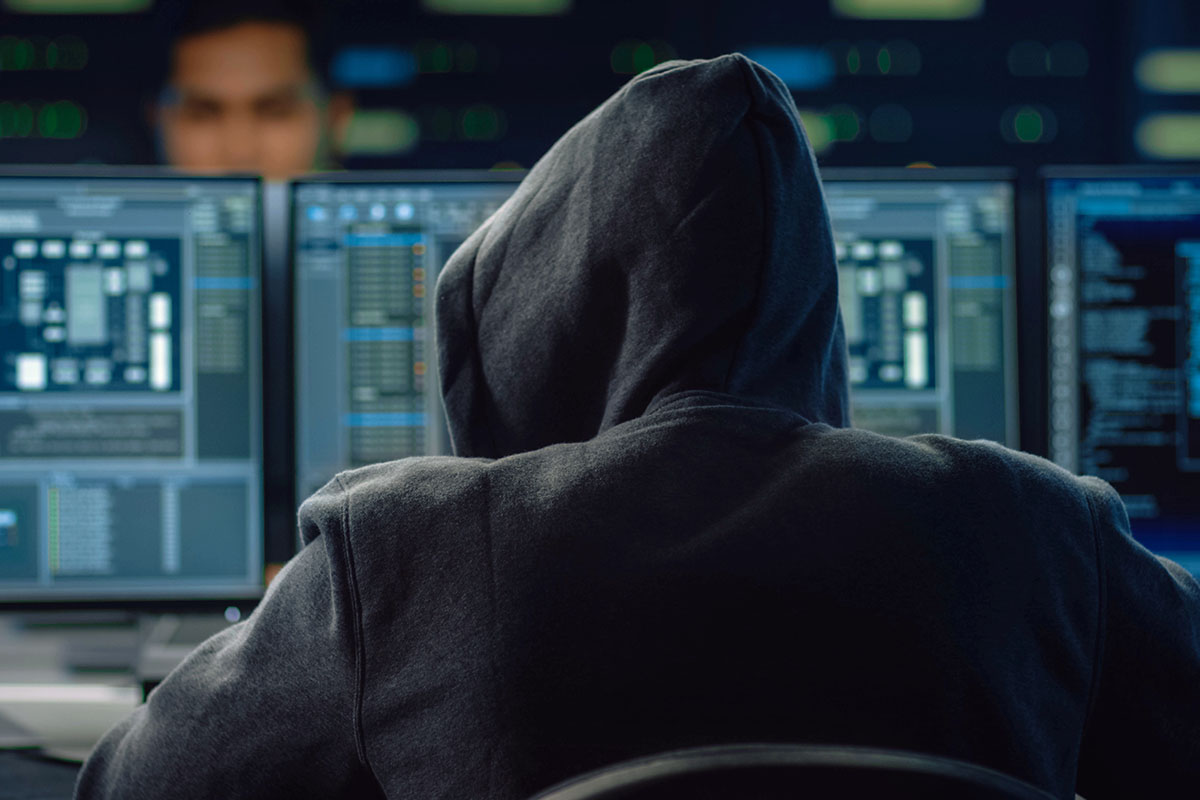 hooded-hacker A person in a dark hoodie seated at a desk, facing multiple computer screens displaying complex data and coding interfaces, with a dimly lit background.