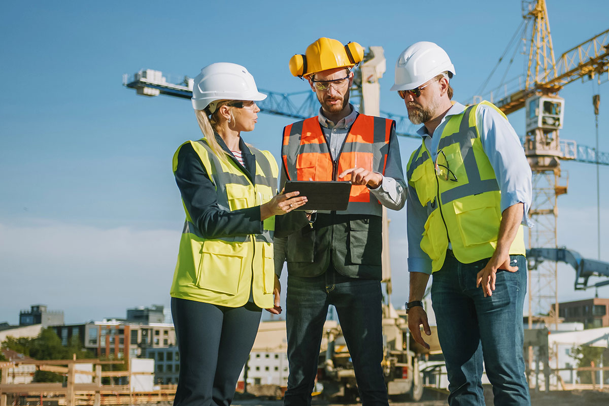 construction03 Three construction workers, two men and one woman, are discussing a project while looking at a tablet. They are wearing hard hats and high-visibility vests. In the background, a construction site with cranes is visible under a clear blue sky.