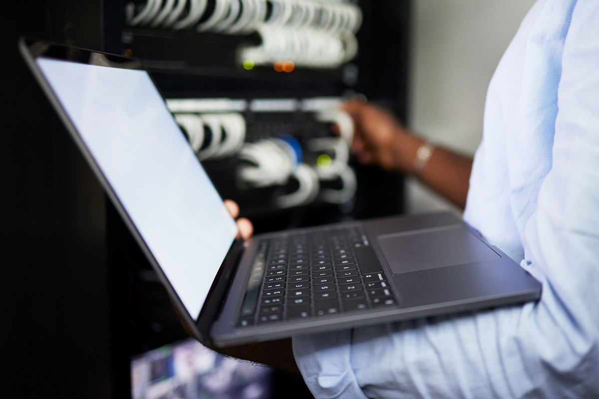 A person holding a laptop with a blank screen while connecting cables in a server rack, with multiple wires and indicators visible in the background.